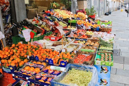 A vibrant street market stall filled with a wide variety of fresh fruits displayed in colorful boxes and crates. The produce includes oranges, apples, pears, grapes, kiwis, and other assorted fruits. Signs with prices and product information are visible.