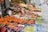 A vibrant street market stall filled with a wide variety of fresh fruits displayed in colorful boxes and crates. The produce includes oranges, apples, pears, grapes, kiwis, and other assorted fruits. Signs with prices and product information are visible.