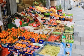 A vibrant street market stall filled with a wide variety of fresh fruits displayed in colorful boxes and crates. The produce includes oranges, apples, pears, grapes, kiwis, and other assorted fruits. Signs with prices and product information are visible.