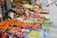 A vibrant street market stall filled with a wide variety of fresh fruits displayed in colorful boxes and crates. The produce includes oranges, apples, pears, grapes, kiwis, and other assorted fruits. Signs with prices and product information are visible.