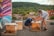 Two people are tending to beehives on a rooftop, with bees visibly active around the hives. One person is wearing a hat and seems to be inspecting one of the hives, while the other is kneeling and adjusting the hive lid. There is a colorful mural on a wall beside them. The background features a view of trees and a hill, with buildings in the mid-ground.