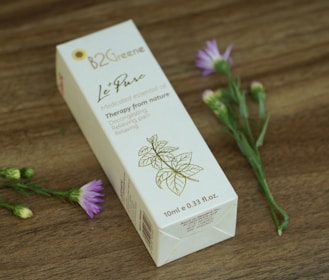 A rectangular box of medicated essential oil is displayed on a wooden surface. The box features elegant typography and a simple floral illustration. Nearby, delicate purple flowers with green stems add a natural touch.