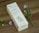 A rectangular box of medicated essential oil is displayed on a wooden surface. The box features elegant typography and a simple floral illustration. Nearby, delicate purple flowers with green stems add a natural touch.