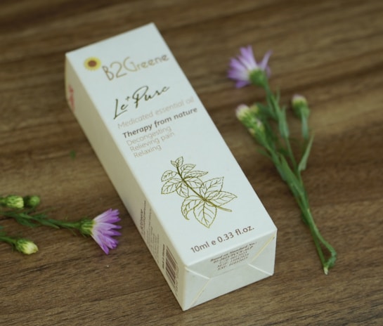A rectangular box of medicated essential oil is displayed on a wooden surface. The box features elegant typography and a simple floral illustration. Nearby, delicate purple flowers with green stems add a natural touch.