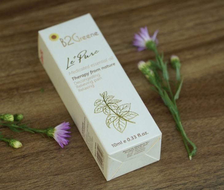 A rectangular box of medicated essential oil is displayed on a wooden surface. The box features elegant typography and a simple floral illustration. Nearby, delicate purple flowers with green stems add a natural touch.