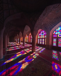An interior view of a beautifully designed architectural space featuring colorful stained glass windows that cast vibrant patterns of light on a richly detailed carpet. The arched ceiling and columns are adorned with intricate patterns, creating a visually captivating scene.