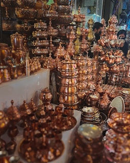An array of intricately designed copperware items such as pots, trays, and utensils, all stacked and displayed in an ornate fashion. The polished copper surfaces reflect light, giving the scene a warm, glowing appearance. Two people are partially visible in the background, likely browsing through the collection.