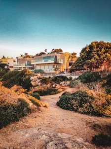 A modern house with large glass windows and balconies is situated on a rocky landscape with various shrubs and trees around. The sky is clear with a warm glow from the sunset, creating an inviting and serene atmosphere.