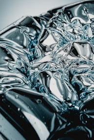 An abstract close-up of a reflective, metallic surface with intricate patterns resembling liquid metal or highly polished chrome. Light plays across the surface, creating dynamic and fluid reflections.