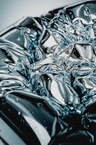 An abstract close-up of a reflective, metallic surface with intricate patterns resembling liquid metal or highly polished chrome. Light plays across the surface, creating dynamic and fluid reflections.