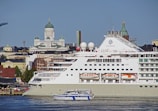 A large cruise ship docks in a harbor with a smaller boat passing by in the foreground. Behind the ship, a cityscape displays historic buildings with domed roofs and ornate architecture, including a prominent cathedral.