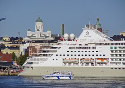 A large cruise ship docks in a harbor with a smaller boat passing by in the foreground. Behind the ship, a cityscape displays historic buildings with domed roofs and ornate architecture, including a prominent cathedral.