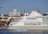 A large cruise ship docks in a harbor with a smaller boat passing by in the foreground. Behind the ship, a cityscape displays historic buildings with domed roofs and ornate architecture, including a prominent cathedral.