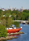 A coastal scene with a large red and black submarine-like vessel positioned on the rocky shore. Surrounding the vessel are lush green trees and a calm body of water reflecting the natural environment. In the background, an urban skyline is visible, including various architectural structures and towers under a clear blue sky.