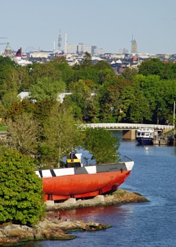 A coastal scene with a large red and black submarine-like vessel positioned on the rocky shore. Surrounding the vessel are lush green trees and a calm body of water reflecting the natural environment. In the background, an urban skyline is visible, including various architectural structures and towers under a clear blue sky.