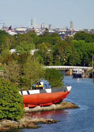 A coastal scene with a large red and black submarine-like vessel positioned on the rocky shore. Surrounding the vessel are lush green trees and a calm body of water reflecting the natural environment. In the background, an urban skyline is visible, including various architectural structures and towers under a clear blue sky.