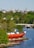 A coastal scene with a large red and black submarine-like vessel positioned on the rocky shore. Surrounding the vessel are lush green trees and a calm body of water reflecting the natural environment. In the background, an urban skyline is visible, including various architectural structures and towers under a clear blue sky.