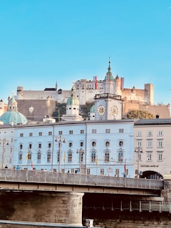 A picturesque cityscape featuring a historic castle perched on a hill, surrounded by brightly colored, classical European architecture. The foreground highlights a bridge with people walking across it, and there is a prominent clock tower and several flags visible.