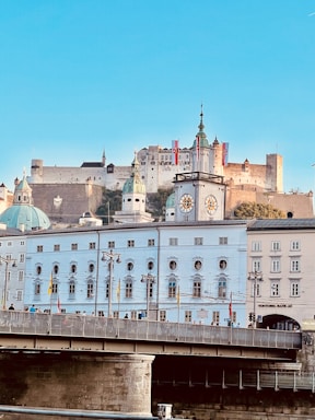 A picturesque cityscape featuring a historic castle perched on a hill, surrounded by brightly colored, classical European architecture. The foreground highlights a bridge with people walking across it, and there is a prominent clock tower and several flags visible.