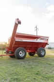 A large, red agricultural grain cart with dual axles and thick tires is positioned on a grassy field. The cart features branding on the side and an extended discharge auger. In the background, there are visible utility poles and a distant building.
