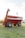 A large, red agricultural grain cart with dual axles and thick tires is positioned on a grassy field. The cart features branding on the side and an extended discharge auger. In the background, there are visible utility poles and a distant building.