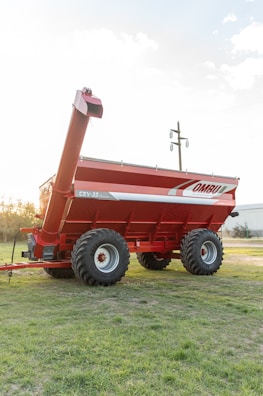 A large, red agricultural grain cart with dual axles and thick tires is positioned on a grassy field. The cart features branding on the side and an extended discharge auger. In the background, there are visible utility poles and a distant building.