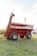 A large, red agricultural grain cart with dual axles and thick tires is positioned on a grassy field. The cart features branding on the side and an extended discharge auger. In the background, there are visible utility poles and a distant building.