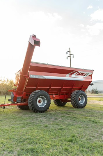 A large, red agricultural grain cart with dual axles and thick tires is positioned on a grassy field. The cart features branding on the side and an extended discharge auger. In the background, there are visible utility poles and a distant building.