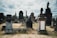 An ancient cemetery with various ornate tombstones and monuments spread across a grassy area. The sky is overcast, adding a somber, atmospheric touch to the scene. Statues and towering structures hint at historical significance, with a variety of textures and architectural styles evident in the grave markers.