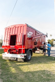 A large red agricultural trailer is parked on grassy ground. It has a shiny metal exterior with visible brand markings. Two people are standing nearby, appearing to evaluate or inspect the trailer. The sun is shining brightly, casting clear shadows, and there are a few palm trees and a partly cloudy sky in the background.