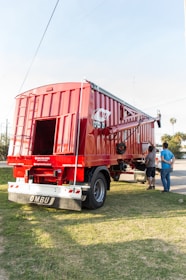 A large red agricultural trailer is parked on grassy ground. It has a shiny metal exterior with visible brand markings. Two people are standing nearby, appearing to evaluate or inspect the trailer. The sun is shining brightly, casting clear shadows, and there are a few palm trees and a partly cloudy sky in the background.