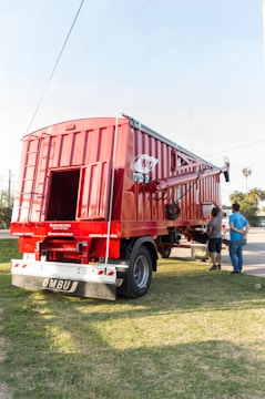 A large red agricultural trailer is parked on grassy ground. It has a shiny metal exterior with visible brand markings. Two people are standing nearby, appearing to evaluate or inspect the trailer. The sun is shining brightly, casting clear shadows, and there are a few palm trees and a partly cloudy sky in the background.