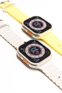 Two smartwatches with rectangular faces are displayed on white and yellow bands. The watches show a detailed watch face with various data points and graphics, including a compass and fitness tracker. The bands have ribbed designs and an orange button is visible on the side of each watch case.