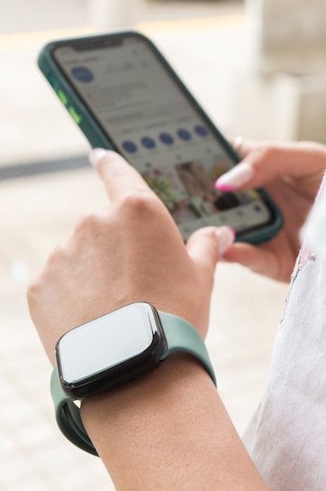A person is using a smartphone with their right hand while wearing a smartwatch with a green strap on their left wrist. The phone screen displays a social media app. The person is indoors in a well-lit area, and their fingernails are painted.
