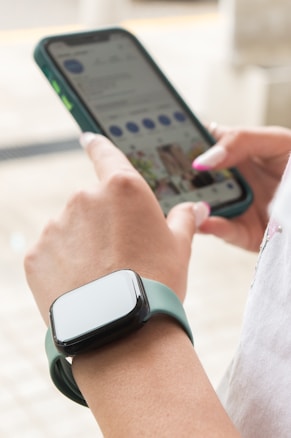 A person is using a smartphone with their right hand while wearing a smartwatch with a green strap on their left wrist. The phone screen displays a social media app. The person is indoors in a well-lit area, and their fingernails are painted.