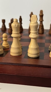 A close-up view of a chessboard featuring several chess pieces. In the foreground, white chess pieces, including a king and queen, are prominently displayed. Brown wooden pieces are visible in the background, slightly out of focus. The board consists of alternating dark and light squares.