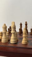 A close-up view of a chessboard displaying several wooden chess pieces in an ongoing game. The pieces are arranged on a classic checkered board, with a mix of light and dark colored pieces.