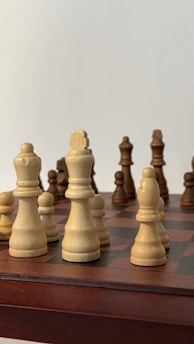 A close-up view of a chessboard displaying several wooden chess pieces in an ongoing game. The pieces are arranged on a classic checkered board, with a mix of light and dark colored pieces.