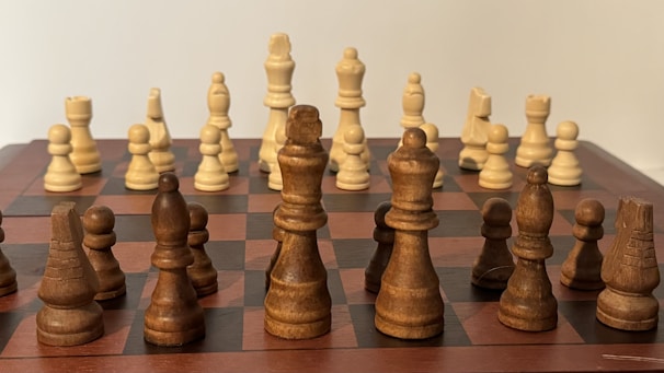 A chessboard with a full set of wooden chess pieces arranged in their starting positions. The pieces are of two contrasting colors, light and dark wood, giving the board a classic and elegant appearance.
