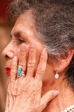 An elderly person with gray hair and wrinkles on their skin. A turquoise ring adorns their finger, pressed gently against their cheek. They are wearing bright red lipstick and pearl earrings.