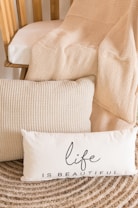 A cozy setup featuring a wooden chair with a beige textured blanket draped over it. Two cushions are placed in front, one with a waffle-knit pattern and another with the phrase 'life is beautiful' printed on it. The surroundings include a woven circular rug that complements the neutral color palette.