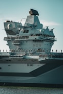 A large naval aircraft carrier with the identifier R09 on its side. The ship has a prominent radar system on top, and several lifeboats and antennas are visible. Numerous crew members stand along the edge, silhouetted against the structure. The vessel is partially submerged in water, and the lighting indicates a clear day.