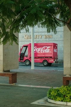 A red Coca-Cola delivery truck is parked in front of a building with the words 'Quality Inn' visible on the wall. The scene is framed by stone pillars and leafy green trees, with a patch of green plants in the foreground.