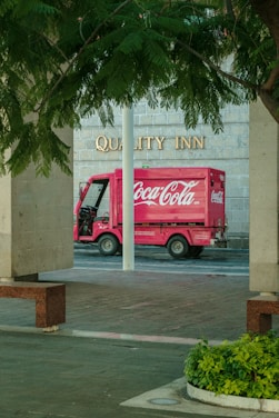 A red Coca-Cola delivery truck is parked in front of a building with the words 'Quality Inn' visible on the wall. The scene is framed by stone pillars and leafy green trees, with a patch of green plants in the foreground.