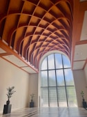 A spacious modern interior features a large, intricate wooden ceiling with a geometric lattice pattern. Tall glass blocks form an expansive arched window, allowing natural light to flood the space. The room is minimally decorated, with two large potted plants on either side.