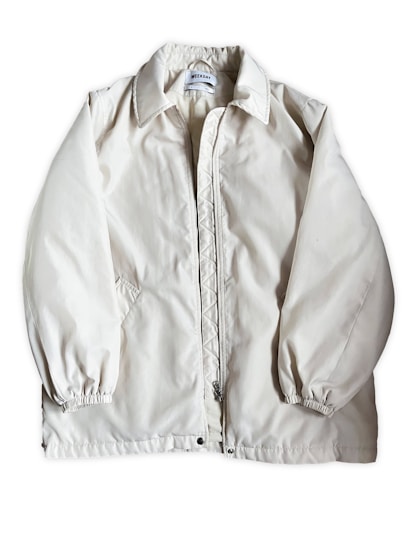 A light beige or off-white jacket with a zipper closure and a collar. The jacket has long sleeves with elastic cuffs and side pockets. The fabric appears to be smooth and slightly shiny. A label is visible on the inside near the collar.