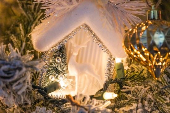 A snowy Christmas scene featuring a small white house adorned with tinsel, a reindeer figurine, and a Christmas tree. Warm fairy lights illuminate the setting, highlighting the delicate textures of the snow-covered branches. A golden, faceted ornament hangs to the side, reflecting the lights.