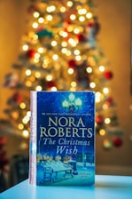 A book titled 'The Christmas Wish' by Nora Roberts is prominently displayed in the foreground, resting on a flat surface. The background features a warmly lit Christmas tree adorned with lights and ornaments, creating a cozy and festive atmosphere.