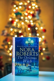 A book titled 'The Christmas Wish' by Nora Roberts is prominently displayed in the foreground, resting on a flat surface. The background features a warmly lit Christmas tree adorned with lights and ornaments, creating a cozy and festive atmosphere.
