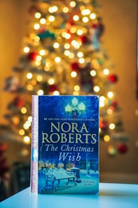 A book titled 'The Christmas Wish' by Nora Roberts is prominently displayed in the foreground, resting on a flat surface. The background features a warmly lit Christmas tree adorned with lights and ornaments, creating a cozy and festive atmosphere.